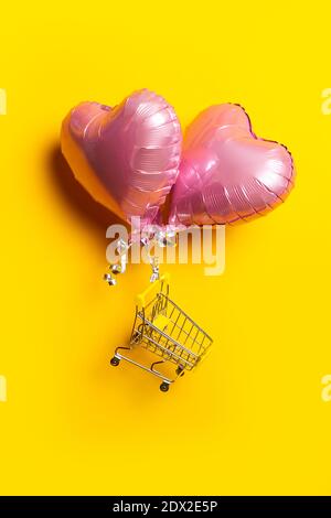 Leerer Einkaufswagen fliegt auf rosa herzförmigen Ballons auf gelbem Hintergrund. Stockfoto