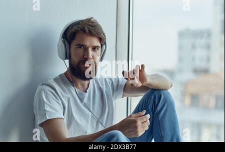 Ein Mann mit Kopfhörern sitzt auf der Fensterbank und hört zu Zur Musik Imitation Fahren eines Autos Lenkrad Studio Stockfoto