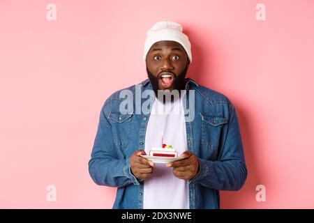 Aufgeregt Black Mann Geburtstag feiern, starrte auf Kamera erstaunt, während Wunschvorstellung auf Geburtstagskuchen mit Kerze, stand über rosa Hintergrund Stockfoto