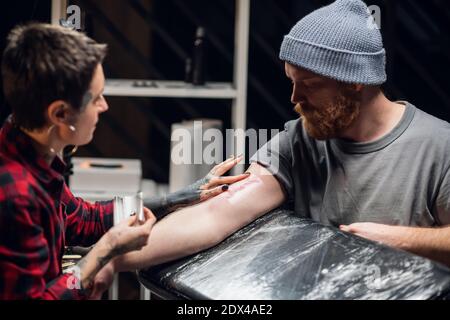 Junge informelle Mädchen Tattoo Künstler setzt einen Verband mit Vaseline Auf der Hand eines jungen Mannes nach erfolgreicher Bewerbung Ein Tattoo auf seiner Hand Stockfoto