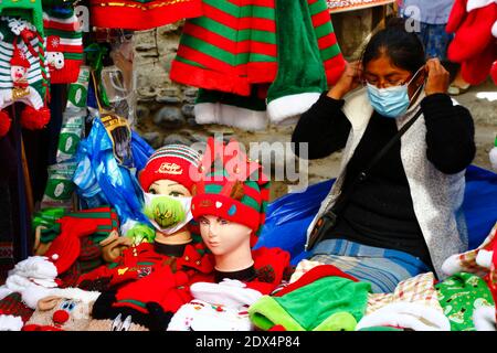 23. Dezember 2020, LA PAZ, BOLIVIEN: Eine Straßenverkäuferin zieht eine Gesichtsmaske an, während sie an ihrem Stand sitzt und Hüte und Gesichtsmasken (darunter eine mit dem Grinch) auf einem Weihnachtsmarkt verkauft. Gesichtsmasken mit Grinch und anderen Weihnachtsthemen sind ein neues Design in den Märkten als Folge der covid 19 Coronavirus Pandemie. Stockfoto