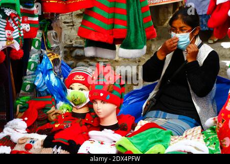 23. Dezember 2020, LA PAZ, BOLIVIEN: Ein Straßenhändler passt ihre Gesichtsmaske an, während er an ihrem Stand sitzt und Hüte und Gesichtsmasken (darunter eine mit dem Grinch) auf einem Weihnachtsmarkt verkauft. Gesichtsmasken mit Grinch und anderen Weihnachtsthemen sind ein neues Design in den Märkten als Folge der covid 19 Coronavirus Pandemie. Stockfoto