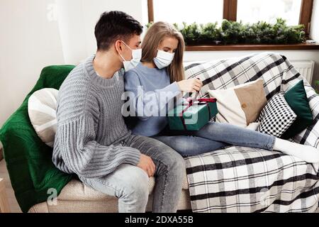 Der junge Mann schenkt seiner Freundin ein Weihnachtsgeschenk, beide tragen Gesichtsmasken Stockfoto