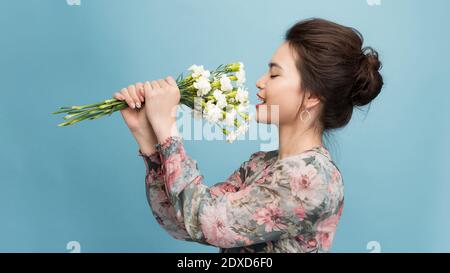 Headshot von hübschen jungen Frau mit zartem Lächeln, gesunde Haut, trägt Strauß von Frühlingsblumen, isoliert auf hellblauem Hintergrund Stockfoto