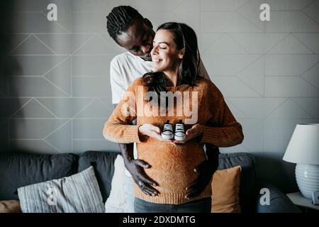 Romantische junge Mann küssen schwangere Frau hält Baby Booties während Zu Hause stehen Stockfoto