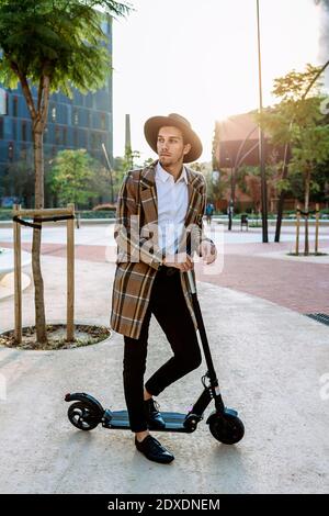 Stilvoller Geschäftsmann mit Hut und Karomuster auf dem Stehen Roller in der Stadt Stockfoto