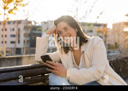 Eine lächelnde Geschäftsfrau, die Smartphone hält, während sie auf der Bank sitzt Stockfoto