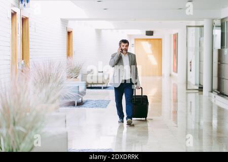 Männlicher Unternehmer am Telefon zieht Gepäck mit Rädern im Hotelkorridor Stockfoto