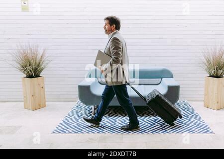 Geschäftsmann zieht Gepäck mit Rädern, während er einen Laptop im Hotel hält Stockfoto