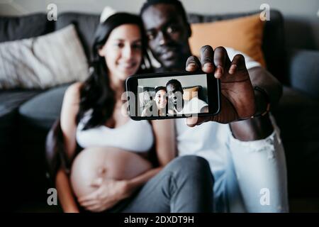 Lächelnder junger Mann mit schwangerer Frau, die zu Hause Selfie nimmt Stockfoto