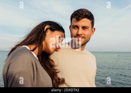 Junge Frau, die am Strand auf der Schulter des Mannes gegen den Himmel lehnt Stockfoto