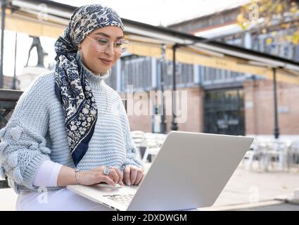 Frau trägt Kopftuch und Brillen arbeiten auf Laptop, während sitzen Im Freien Stockfoto