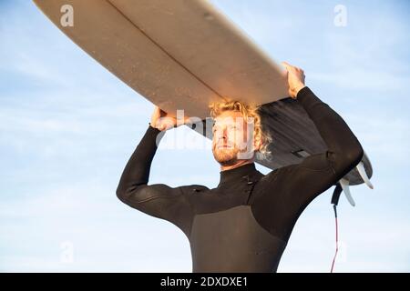 Blonde männliche Surfer trägt Surfbrett über Kopf am Strand gegen Himmel Stockfoto