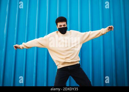 Sorgloser Mann mit Gesichtsmaske, der mit ausgestreckten Armen springt Blaue Wand Stockfoto