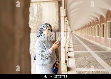 Junge Frau trägt Kopftuch mit Handy, während sich auf Säule Stockfoto