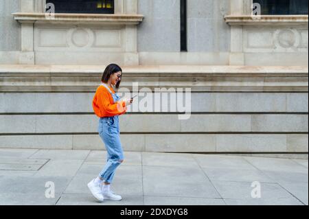 Eine glückliche europäische Frau, die einen orangefarbenen Pullover trägt, geht hinunter Die Straße mit ihrem Handy in den Händen und Lächelnd, als sie die Kamera anschaut.Mad Stockfoto