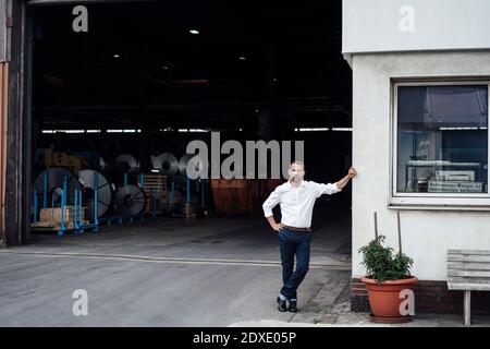 Männlicher Besitzer mit der Hand an der Hüfte, während er sich an der Hütte lehnte Steht gegen den Eingang der Fabrik Stockfoto
