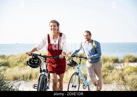 Lächelnder Mann und Frau, die am Strand mit Fahrrädern spazieren Stockfoto