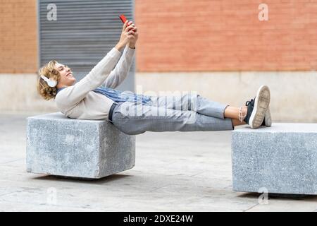 Lächelnder Mann mit Kopfhörern, der Selfie auf Beton aufnahm Bank in der Stadt Stockfoto