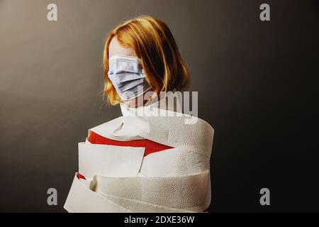 Teenager-Mädchen in Toilettenpapier eingewickelt und Gesicht mit bedeckt Schützende Gesichtsmaske steht vor grauem Hintergrund Stockfoto