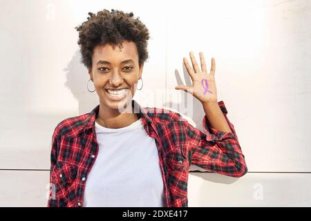 Fröhliche junge Frau mit lila Bewusstsein Symbol auf Handfläche gegen Weiße Wand während des Womens Day Stockfoto