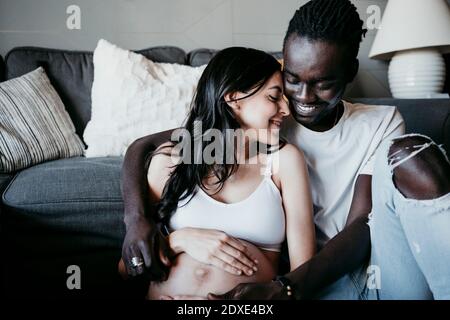 Lächelnder junger Mann mit schwangerer Frau, die im Wohnzimmer sitzt Zu Hause Stockfoto