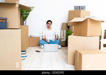 Junger Mann, der Kaffee trinkt, während er auf dem Boden sitzt, umgeben von Boxen in der neuen Wohnung Stockfoto