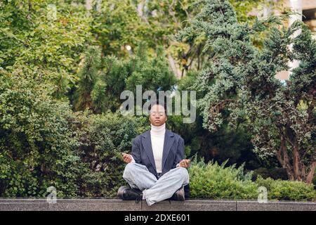 Junge Geschäftsfrau, die Yoga macht, während sie auf einer Stützmauer sitzt Anlagen Stockfoto