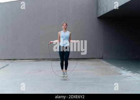 Junge Sportlerin, die gegen die Wand steht und dabei Seil hüpft Stockfoto