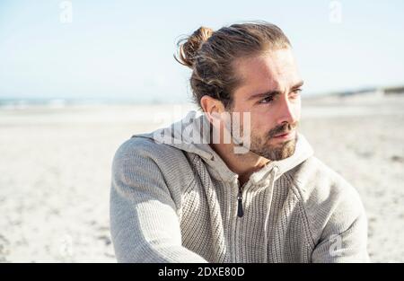 Junger Mann mit Kapuzenhemd, der beim Sitzen wegschaut Strand an sonnigen Tagen Stockfoto