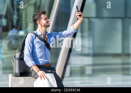 Lächelnder Geschäftsmann, der Selfie über das Mobiltelefon nimmt, während er im Freien sitzt Stockfoto