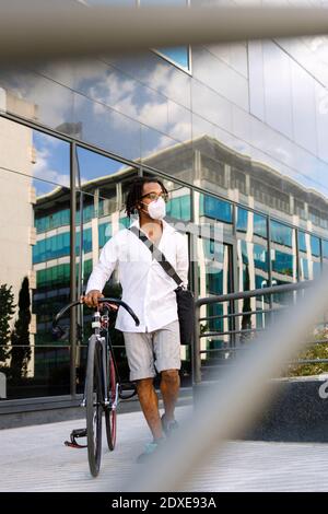 Mann mit Gesichtsmaske, der mit dem Fahrrad gegen ein modernes Gebäude läuft Während der Covid-19 in der Stadt Stockfoto