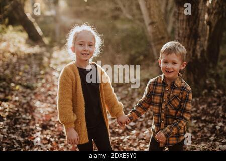 Bruder und Schwester lächeln, während sie die Hände am Wald halten Stockfoto