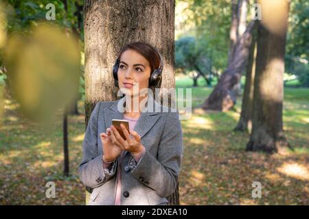 Durchdachte Geschäftsfrau, die Musik hört, während sie Smartphone im Park hält Im Herbst Stockfoto