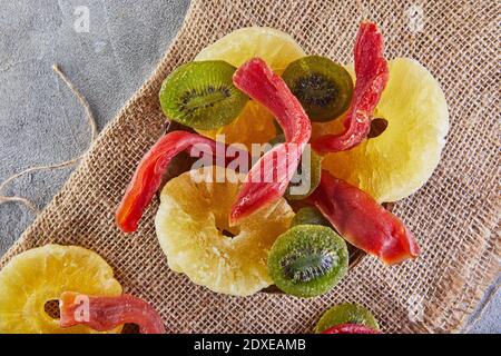 Getrocknete Früchte: Gelbe kandierte Ananasringe, rote Papaya und grüne Kiwi in einem Holzteller auf Sackleinen. Nahaufnahme Stockfoto