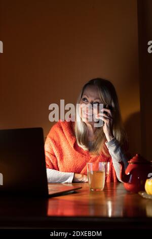 Ältere Frau mit Laptop, die im Sitzen auf dem Mobiltelefon spricht Zu Hause Stockfoto