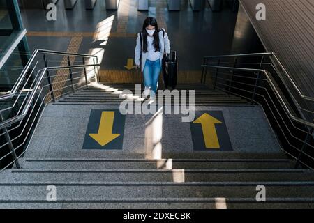 Frau, die Gepäck trägt, während sie sich auf der Treppe bei der Eisenbahn aufbewegt Station während einer Pandemie Stockfoto
