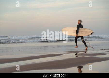 Surfer läuft mit Surfbrett in Richtung Wellen im Meer während Sonnenuntergang Stockfoto