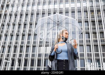 Weibliche professionelle mit Regenschirm hält Einweg-Tasse gegen Gebäude in Stadt während der COVID-19 Stockfoto