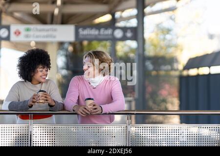 Glückliche multiethnische Freunde halten wiederverwendbare Tassen, während sie sich auf Geländer stützen Am Bahnhof Stockfoto