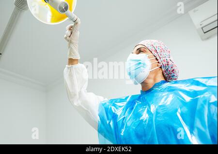 Zahnarzt trägt schützende Arbeitskleidung Einstellung elektrische Lampe während des Stehens an Klinik während Covid-19 Stockfoto