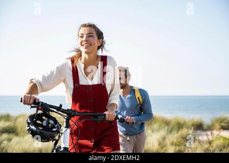 Lächelnde Freundin hält Fahrräder, während sie mit ihrem Freund gegen das Meer läuft Stockfoto