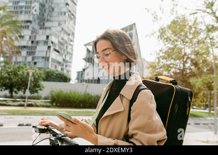 Lächelnde Frau mit Instrumentenkoffer mit Mobiltelefon im Stehen Mit Elektro-Roller in der Stadt Stockfoto