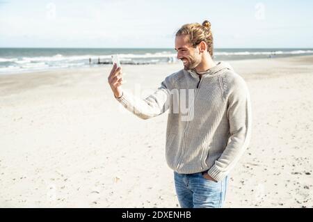 Junger Mann lächelt, während Selfie über Handy zu nehmen Strand Stockfoto