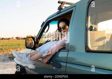 Überraschte junge Frau, die mit den Füßen auf dem Auto wegschaut Fenster während der Fahrt bei Sonnenuntergang Stockfoto