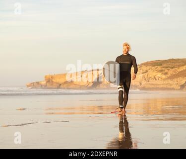 Männlicher Surfer mit Surfbrett, der am Strand gegen den Himmel läuft Stockfoto