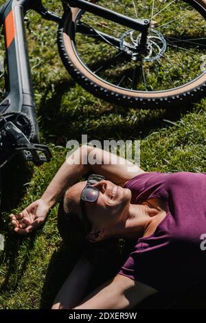 Lächelnde Frau ruht sich aus, während sie mit dem Mountainbike auf dem Gras liegt Im Park Stockfoto