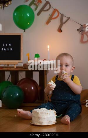 Hungriger Junge essen Kuchen von seinem ersten Geburtstag, während Sitzen gegen Geburtstag Dekoration zu Hause Stockfoto