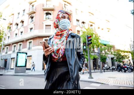 Muslimische Frau in Hijab hält Handy zu Fuß auf der Straße In der Stadt während der COVID-19 Stockfoto