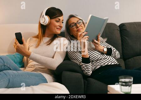 Mutter zeigt der Tochter ein Buch mit dem Handy während sie sitzt Auf dem Sofa zu Hause Stockfoto